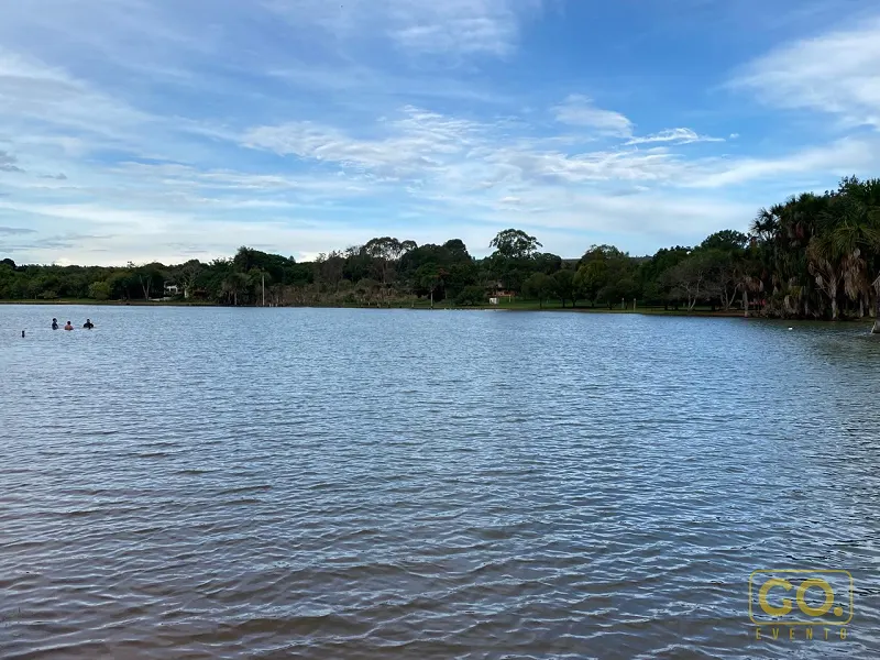 Lago de águas calmas cercado por vegetação densa e palmeiras, com duas pessoas nadando ao longe sob céu azul com nuvens.