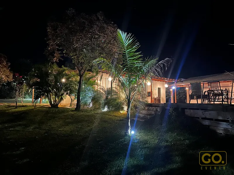 Casa de campo iluminada à noite, com gramado, palmeiras e deck de madeira com mesa e cadeiras ao lado.