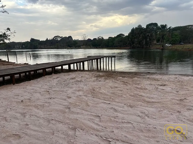Praia de areia às margens de um lago, com deck de madeira avançando sobre a água e árvores ao fundo sob céu nublado.