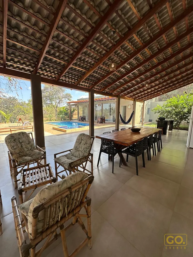 Varanda coberta com telhado de madeira, mesa rústica e cadeiras de bambu, com piscina e rede ao fundo.