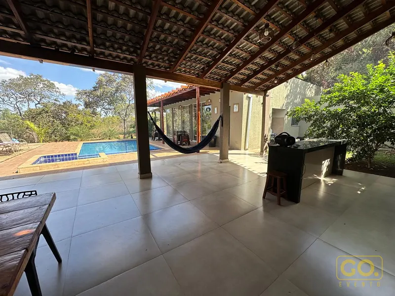 Área coberta com piso de cerâmica clara, rede pendurada e piscina ao fundo, sob sol forte e vegetação ao redor.