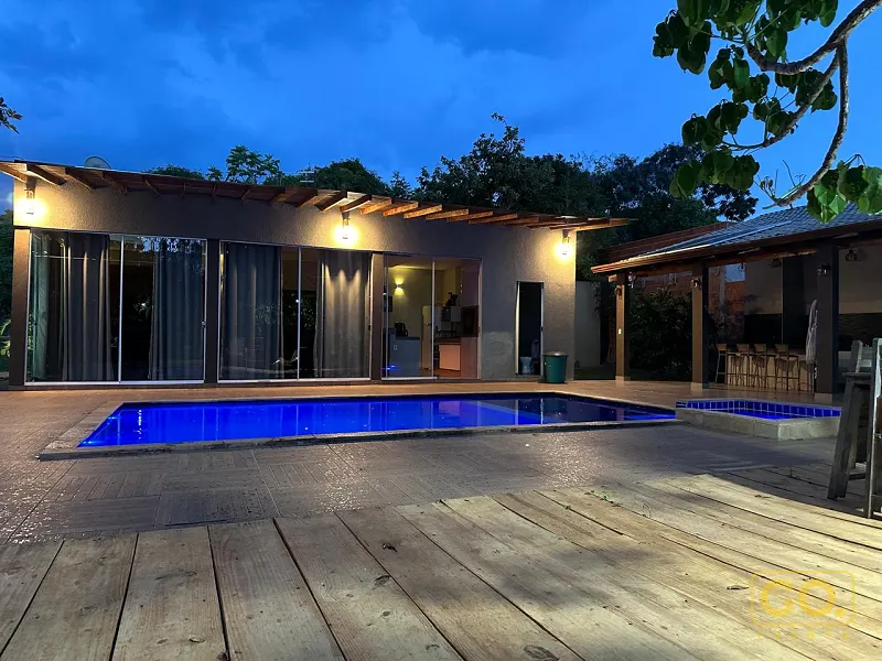 Piscina iluminada em azul ao anoitecer, ao lado de deck de madeira e de uma casa com portas de vidro e área coberta com bar.
