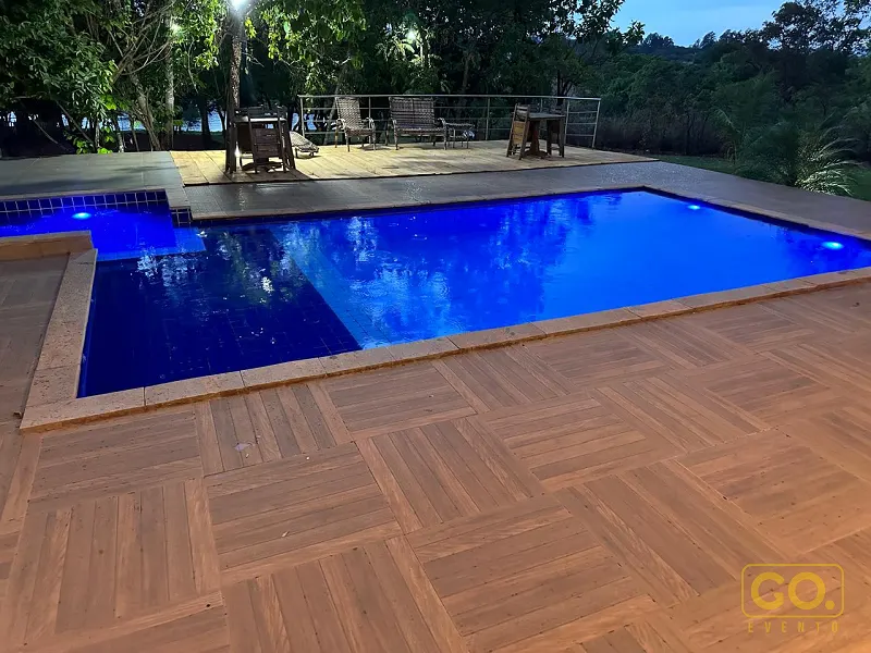 Piscina retangular iluminada em azul à noite, com deck de madeira e área com cadeiras e guarda-corpo ao fundo.