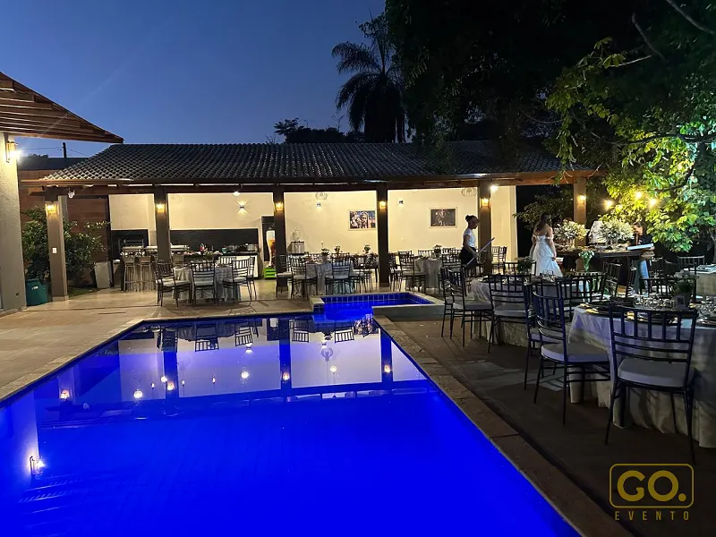 Piscina iluminada em azul à noite, com mesas redondas postas e cadeiras pretas ao redor em área com pergolado.