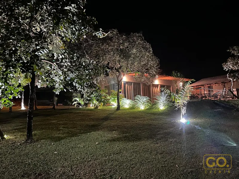 Jardim gramado iluminado à noite, com árvores, palmeiras e uma casa de telhado de telha ao fundo.