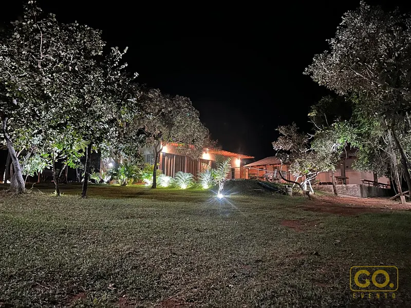 Gramado e árvores iluminadas à noite, com luminárias de solo destacando o jardim e as construções ao fundo.