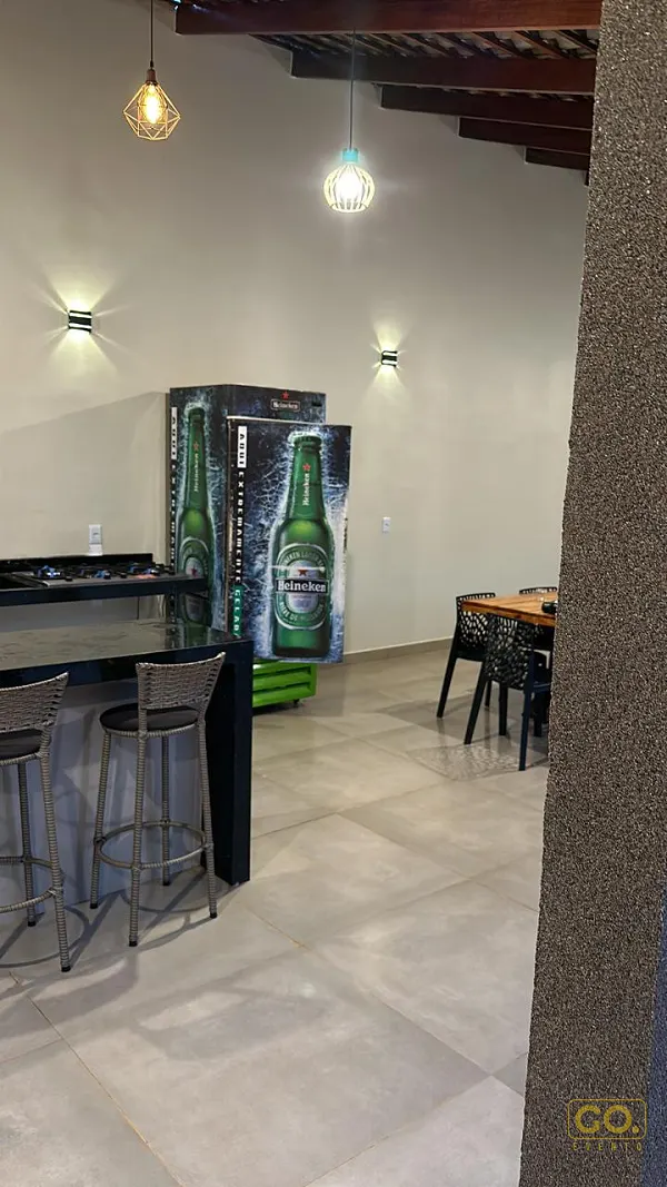 Balcão preto com cooktop e banquetas de vime em área coberta, com luminárias pendentes e mesa de madeira com cadeiras ao fundo.