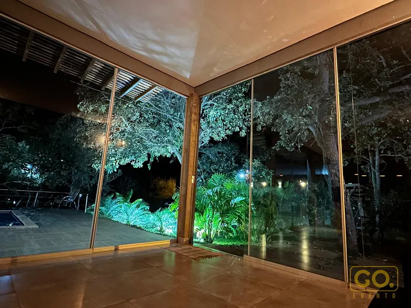Varanda coberta com paredes de vidro à noite, com vista para jardim iluminado, árvores e piscina ao fundo.