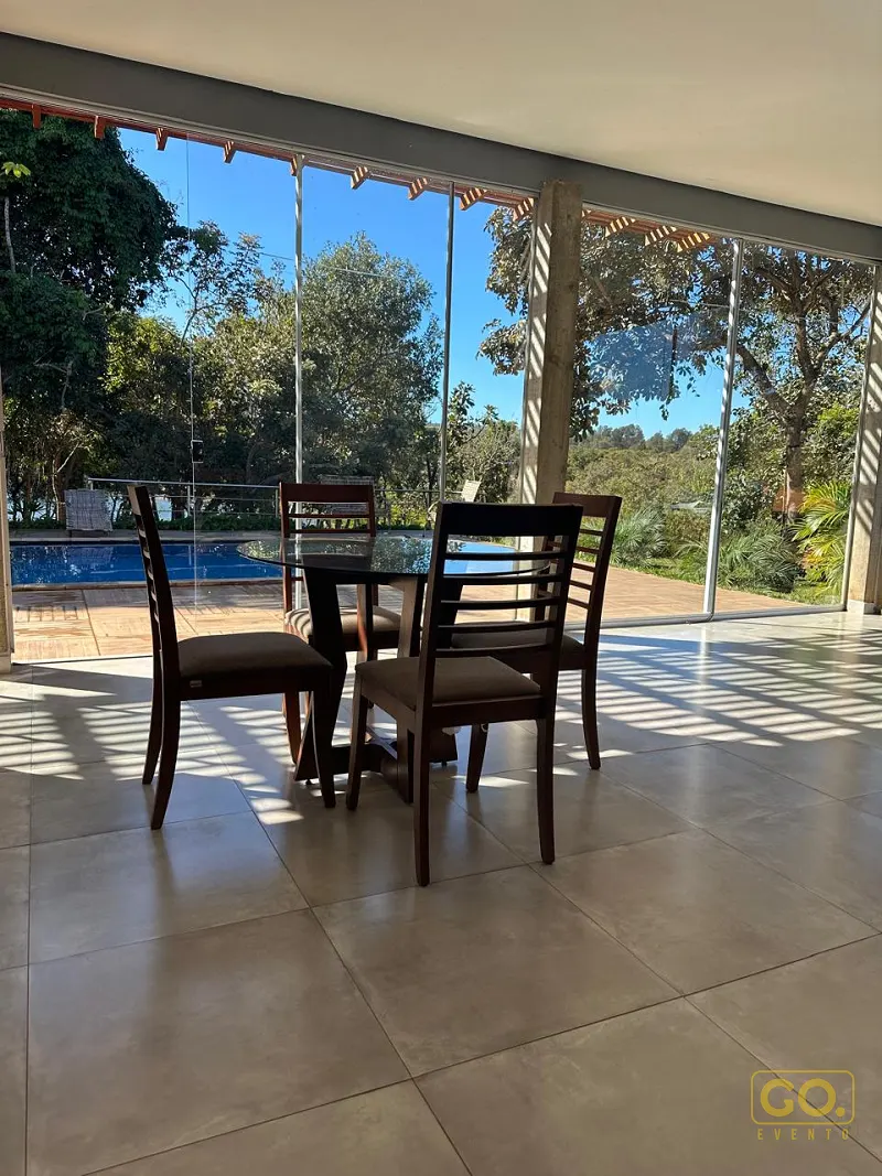 Salão envidraçado com mesa redonda e quatro cadeiras de madeira, com vista para piscina e árvores ao fundo.