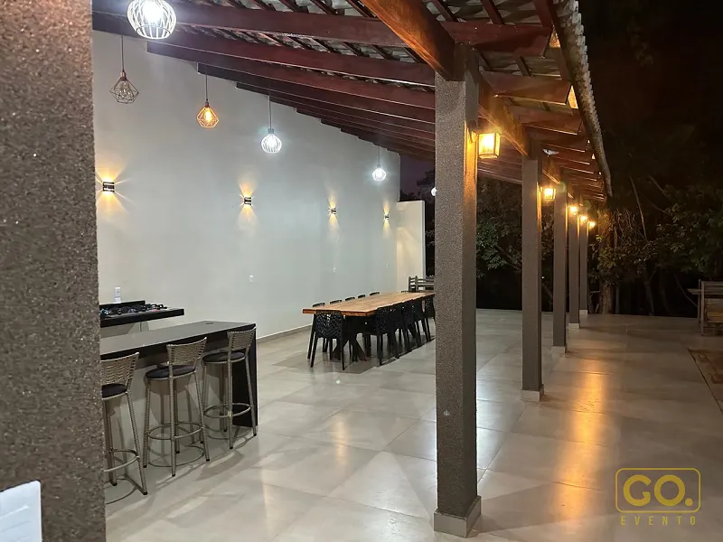 Área coberta com pérgola de madeira, colunas revestidas, luminárias pendentes e mesa comprida ao fundo, iluminada à noite.