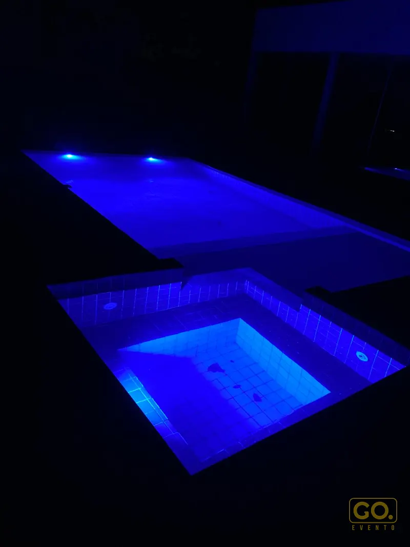 Piscina iluminada por luz azul à noite, com uma piscina menor de azulejos ao lado, sem pessoas presentes.