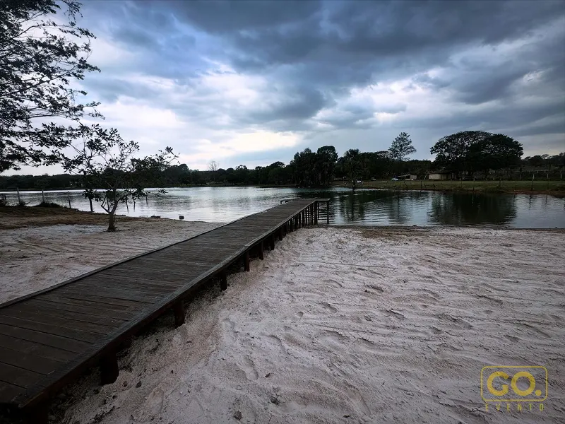Píer de madeira que avança sobre a areia até um lago calmo, sob céu nublado e com árvores ao fundo.