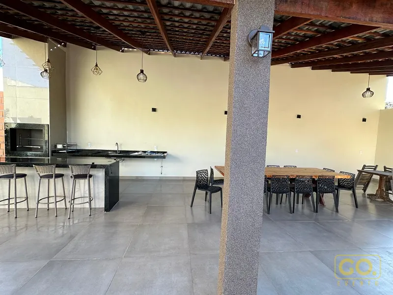 Área gourmet coberta com churrasqueira embutida, balcão de granito com banquetas e mesa de madeira com cadeiras pretas ao fundo.