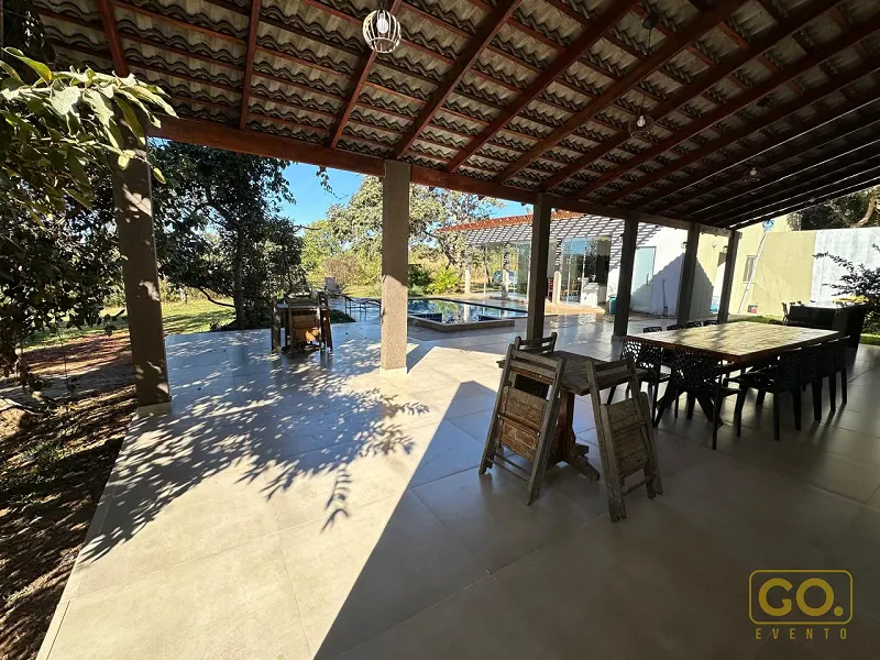 Área externa coberta com telhado de telha, mesa de madeira com cadeiras, churrasqueira e piscina ao fundo.
