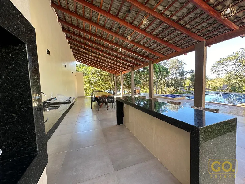 Área coberta com bancada de granito preto e pia, com piscina e vegetação ao fundo em dia de sol.
