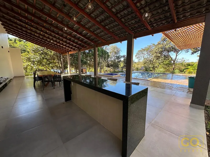Área coberta com bancada de granito preto, mesa de jantar ao fundo e piscina com árvores ao redor, vista pelo pergolado de madeira.