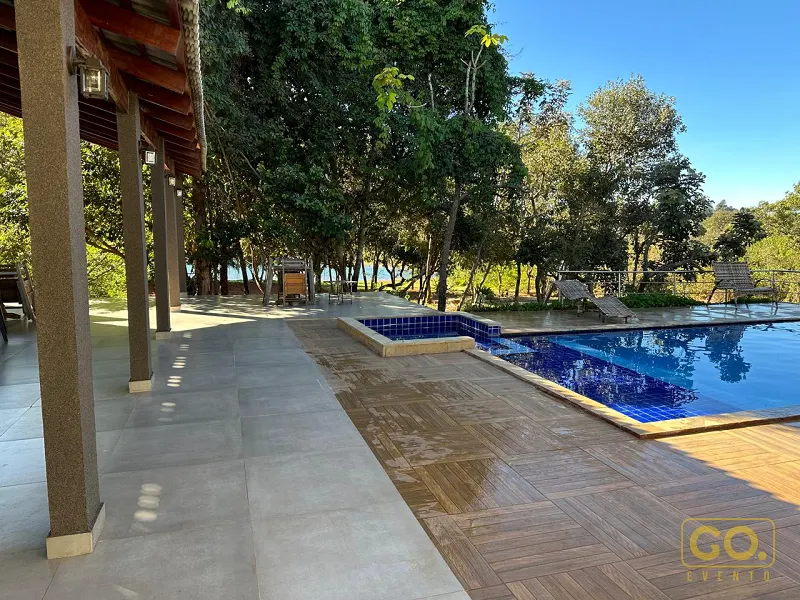 Piscina de azulejos azuis com deck de madeira ao lado de área coberta com pilares, cercada por árvores e vista para a água.