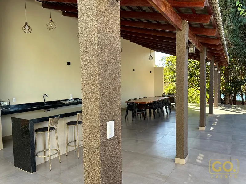 Área gourmet coberta com bancada de granito e pia, banquetas altas e mesa comprida de madeira ao fundo, sob vigas de madeira.
