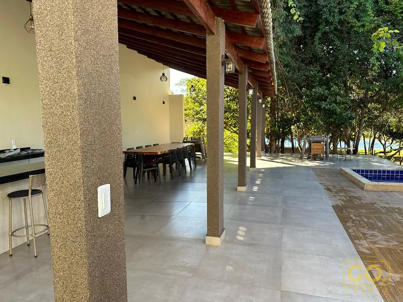 Varanda coberta com mesa de madeira comprida, balcão com banquetas, piscina à direita e vegetação ao fundo.