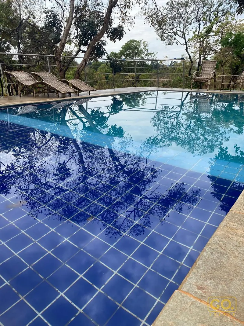 Piscina de azulejos azuis refletindo árvores, com espreguiçadeiras de palha ao fundo e borda de pedra em área externa.