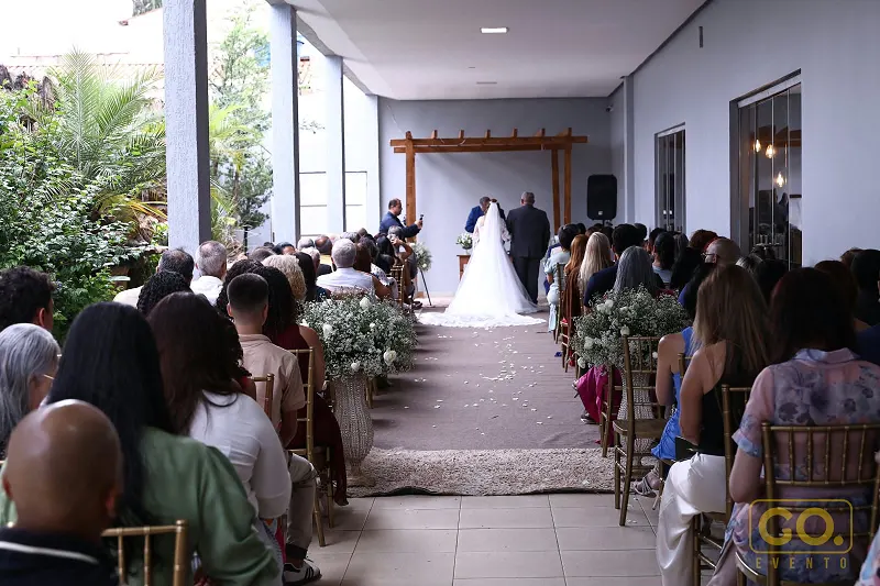Cerimônia de casamento em espaço coberto, com noivos no altar sob arco de madeira e convidados sentados nas fileiras laterais.
