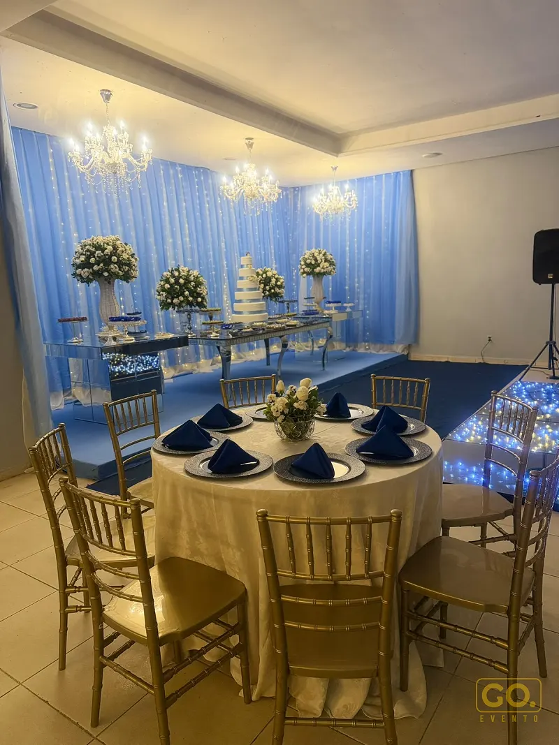 Salão decorado em azul e branco com mesa redonda posta, bolo de vários andares ao fundo e lustres de cristal no teto.