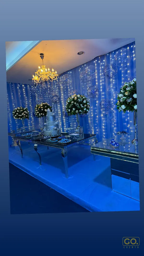 Salão decorado em azul com cortina de luzes, lustre de cristal, bolo de andares e mesas espelhadas com flores brancas.
