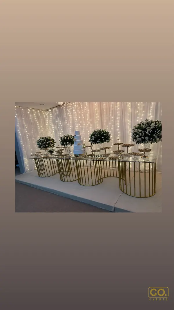 Mesa de vidro sobre bases douradas com arranjos de flores, doces e bolo branco, diante de cortina com luzes de fada.