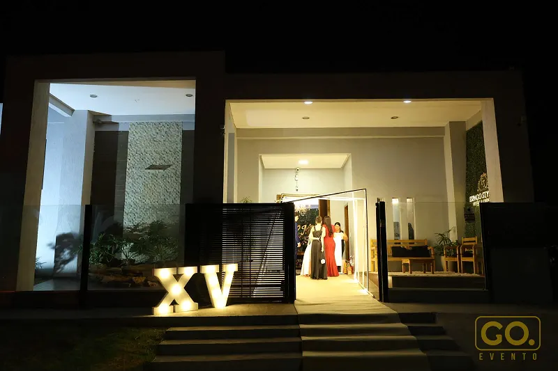Fachada noturna de espaço com letras XV iluminadas na entrada, portão de ripas escuras e grandes painéis de vidro.