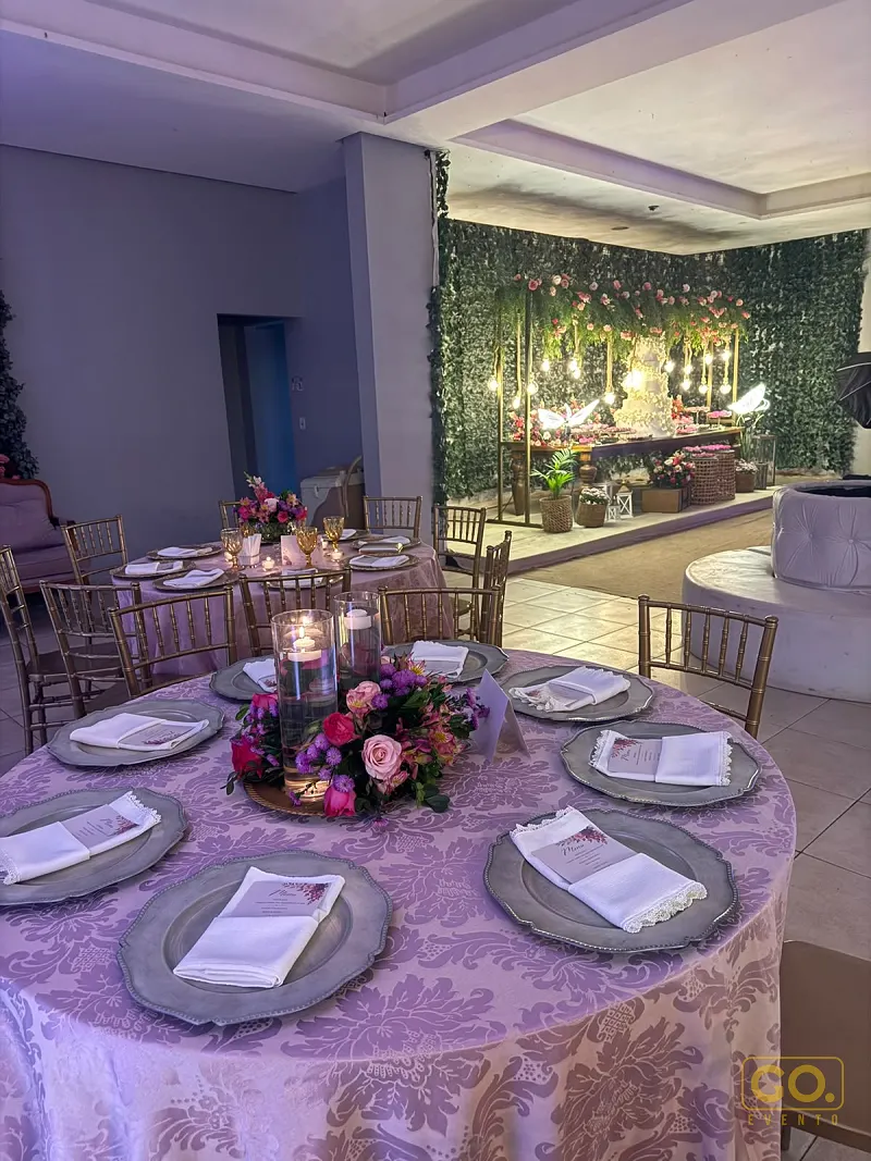 Salão decorado com mesas redondas postas, toalhas lilás de damasco e painel verde com flores ao fundo, sem convidados.
