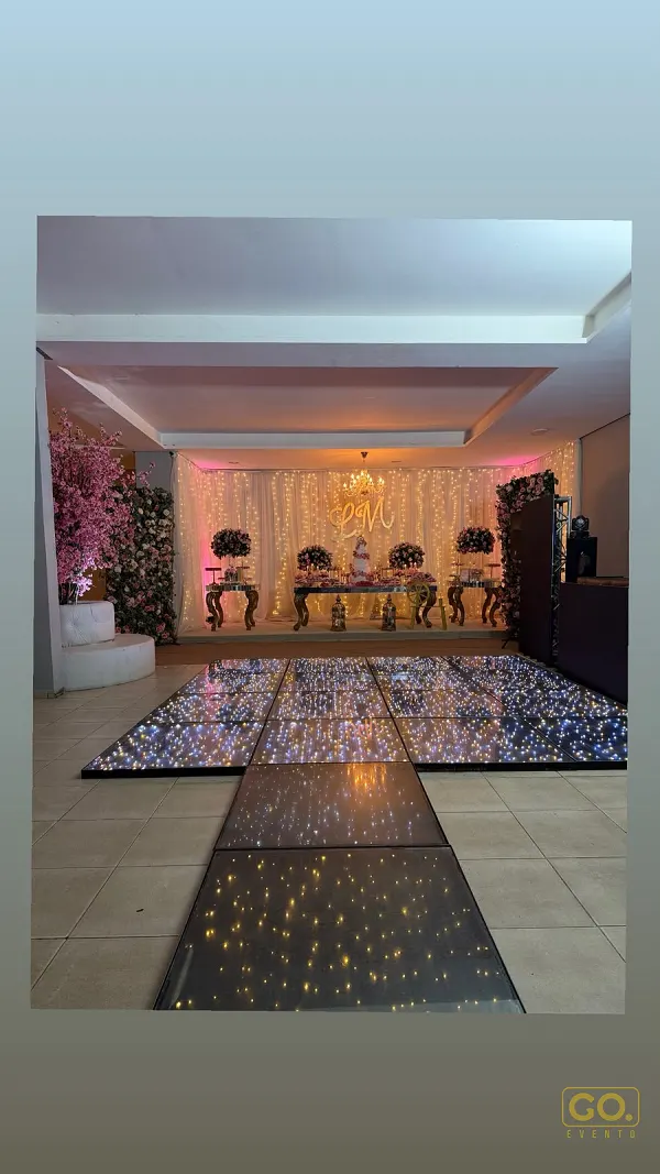 Salão decorado com cortina de luzes, mesa de doces ao fundo e pista de dança com piso de LED cintilante, sem convidados presentes.