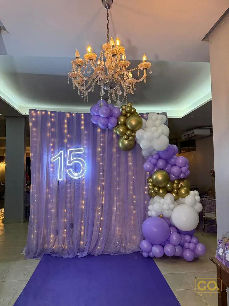 Entrada decorada com cortina lilás iluminada, letreiro de neon com o número 15 e arco de balões roxos, dourados e brancos.