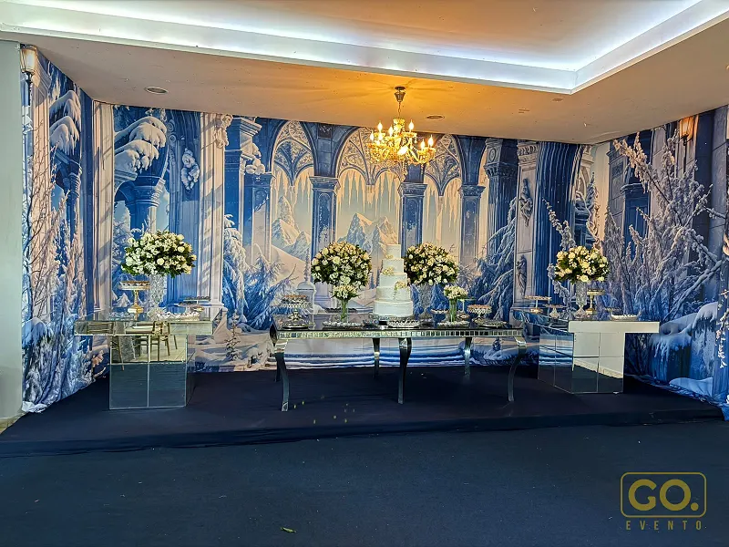 Mesa de bolo decorada com arranjos de flores brancas em frente a painel com mural de inverno em tons de azul e branco.