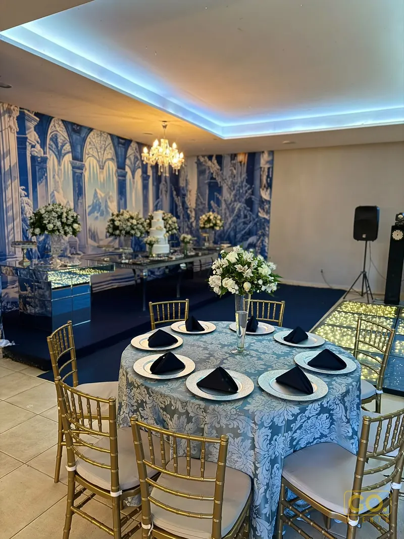 Salão decorado em azul e branco com mesa redonda posta, cadeiras douradas e mesa de bolo ao fundo sob mural pintado.