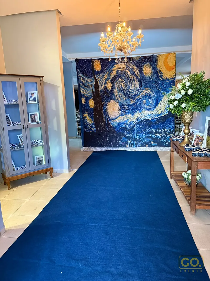 Hall decorado com painel da obra A Noite Estrelada, tapete azul no chão, lustre de cristal e mesa com lembrancinhas azuis.