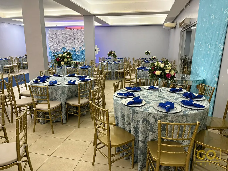 Salão de festas decorado com mesas redondas de toalha azul-clara, guardanapos azuis e cadeiras douradas, sem convidados.