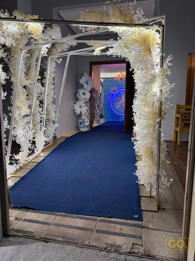 Entrada decorada com arco de flores brancas e fitas de LED, com tapete azul levando a um salão iluminado ao fundo.