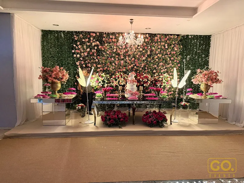 Mesa de doces e bolo de vários andares em frente a uma parede coberta de flores cor-de-rosa, com lustre de cristal ao alto.