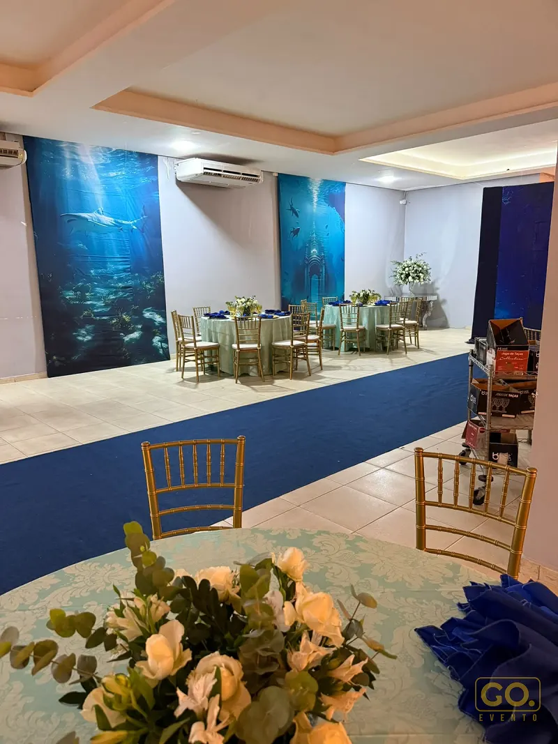 Salão decorado com tema marinho, mesas redondas postas com toalhas verde-menta e cadeiras douradas, sem convidados.