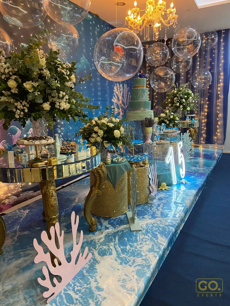 Salão decorado com tema de fundo do mar, com bolhas transparentes penduradas, bolo turquesa de três andares e mesa de doces espelhada e dourada.