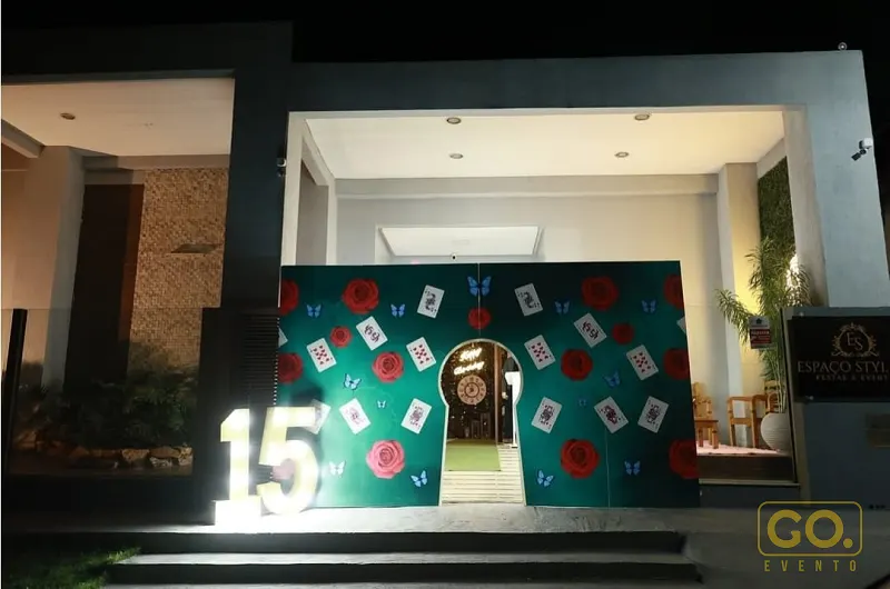 Fachada noturna decorada para festa de 15 anos, com painel verde, rosas vermelhas, cartas, borboletas e número 15 iluminado.