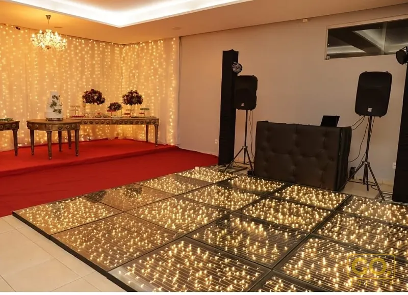 Salão decorado com cortina de luzes, pista de dança iluminada, mesa com bolo e flores e equipamento de DJ.
