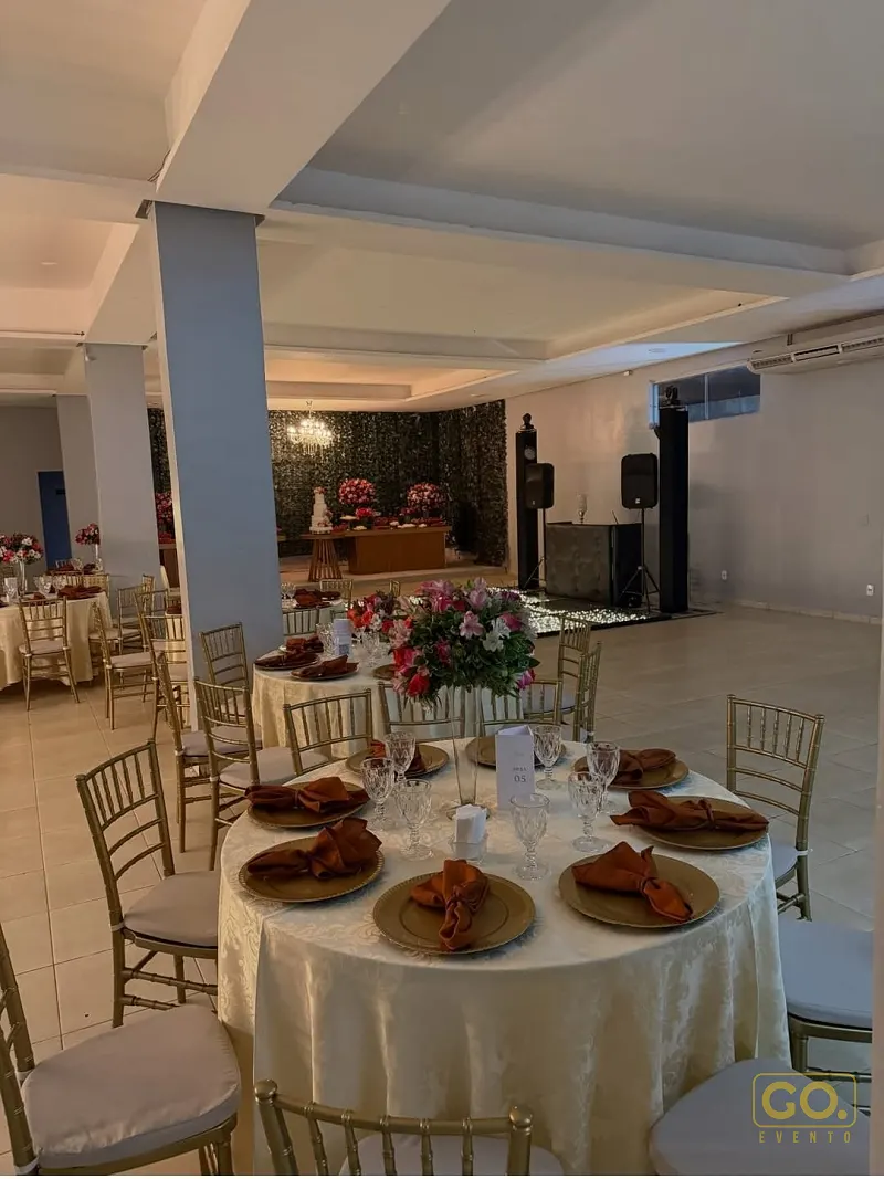 Salão com mesas redondas postas com toalhas creme, cadeiras douradas e arranjos de flores rosa e terracota ao fundo.