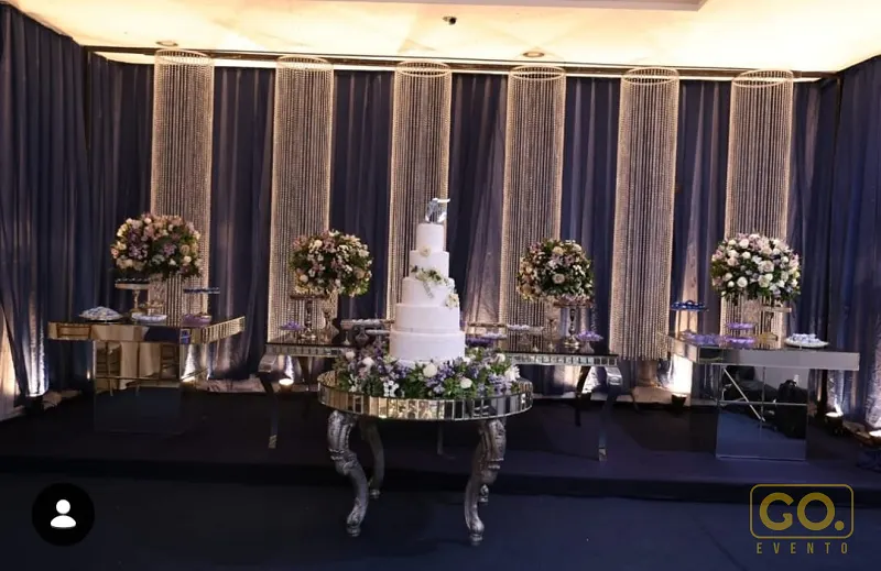 Mesa de bolo de casamento de vários andares coberta de flores, entre cortinas de cristal, espelhos e tecidos azul-marinho iluminados