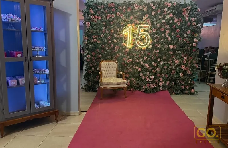 Hall decorado com painel de flores rosas, letreiro de neon com o número 15, tapete vermelho e poltrona estofada ao centro.