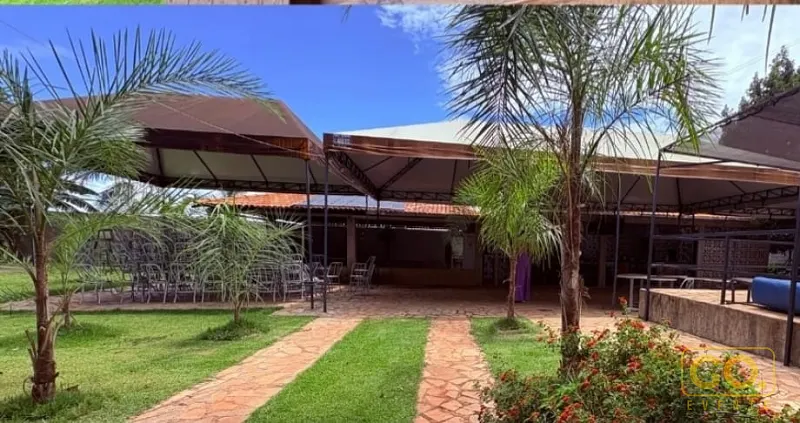 Área externa de chácara com cobertura de lona, cadeiras empilhadas, gramado, palmeiras e caminho de pedra até o salão ao fundo.