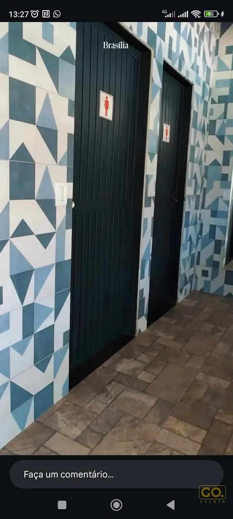Corredor com azulejos geométricos azuis e brancos e duas portas de madeira escura, sinalizadas como banheiros feminino e masculino.
