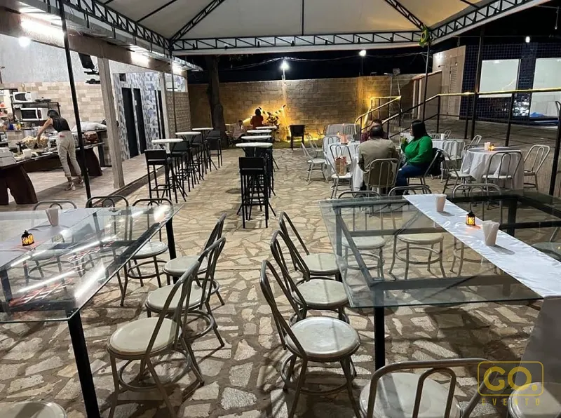 Área externa à noite com piso de pedra, mesas de vidro com toalha branca, cadeiras e um balcão de preparo ao fundo.