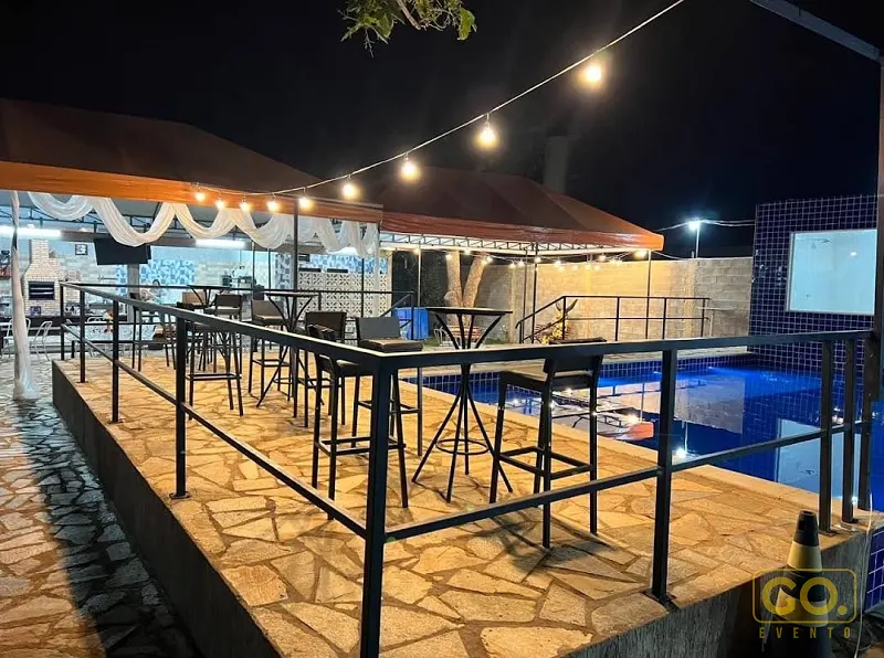 Deck de pedra à noite com mesas e banquetas altas ao lado de uma piscina iluminada por cordões de lâmpadas.