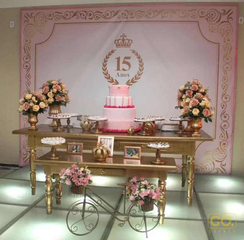 Mesa dourada com bolo rosa de três andares, painel com coroa e 15 Anos, arranjos de flores e petiscos em decoração de festa.